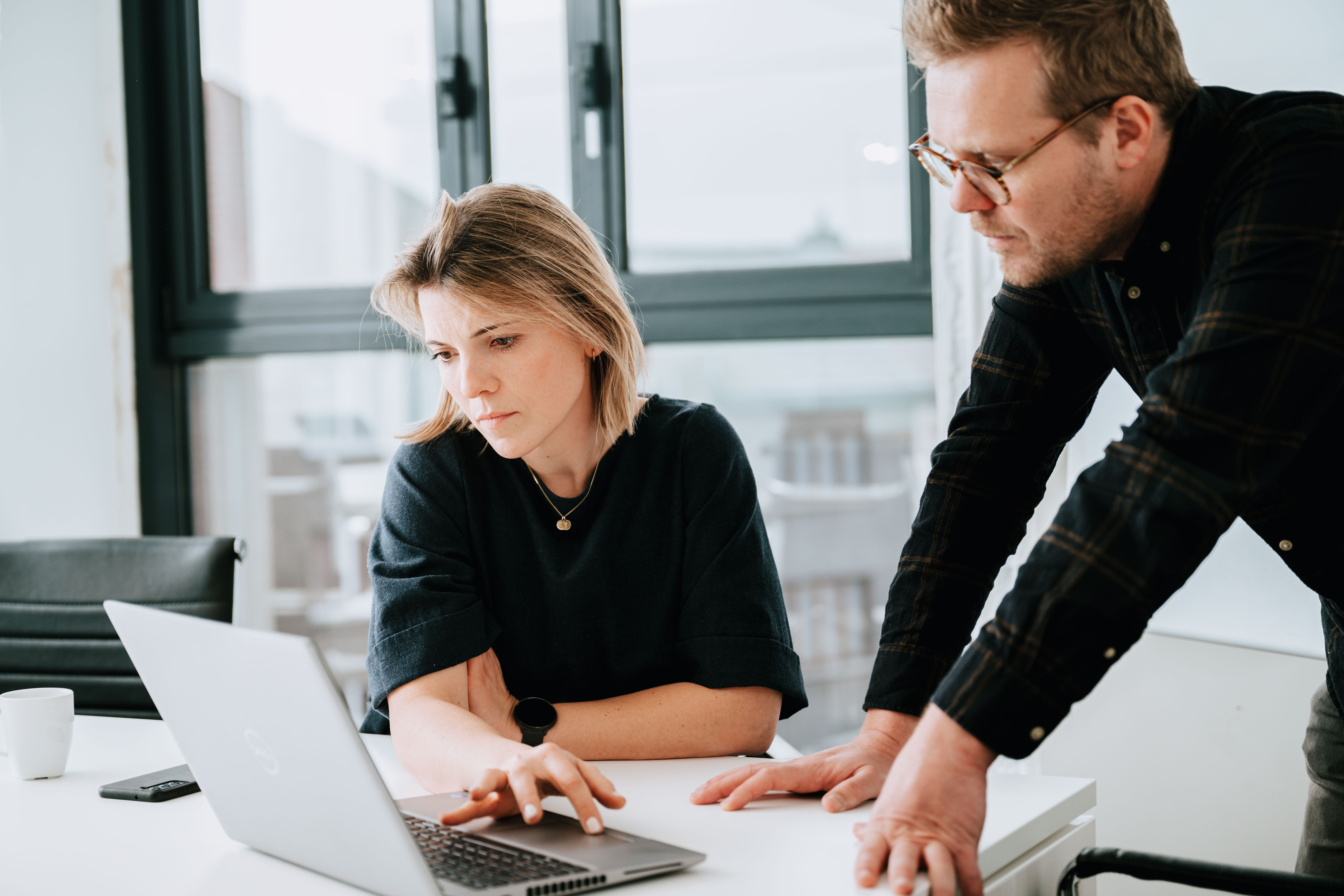 Two people watching a laptop screen, one is sitting down while the other leans over the desk