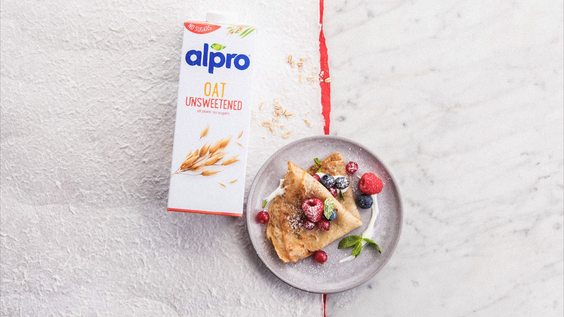 Some pancakes on a plate accompanied by fresh fruit and an Alpro oat unsweetened bottle of milk 